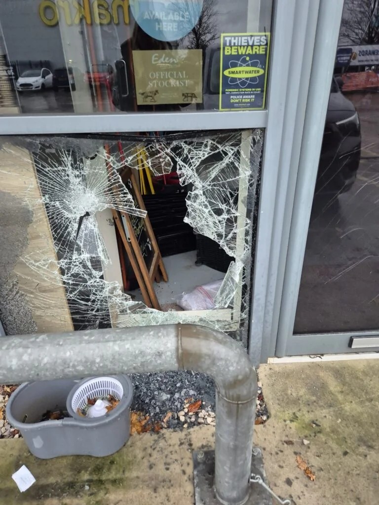 Smashed low-level glazed panel in an aluminium shopfront on Deeside Industrial Estate after a break-in, with the shop interior visible through the broken glass