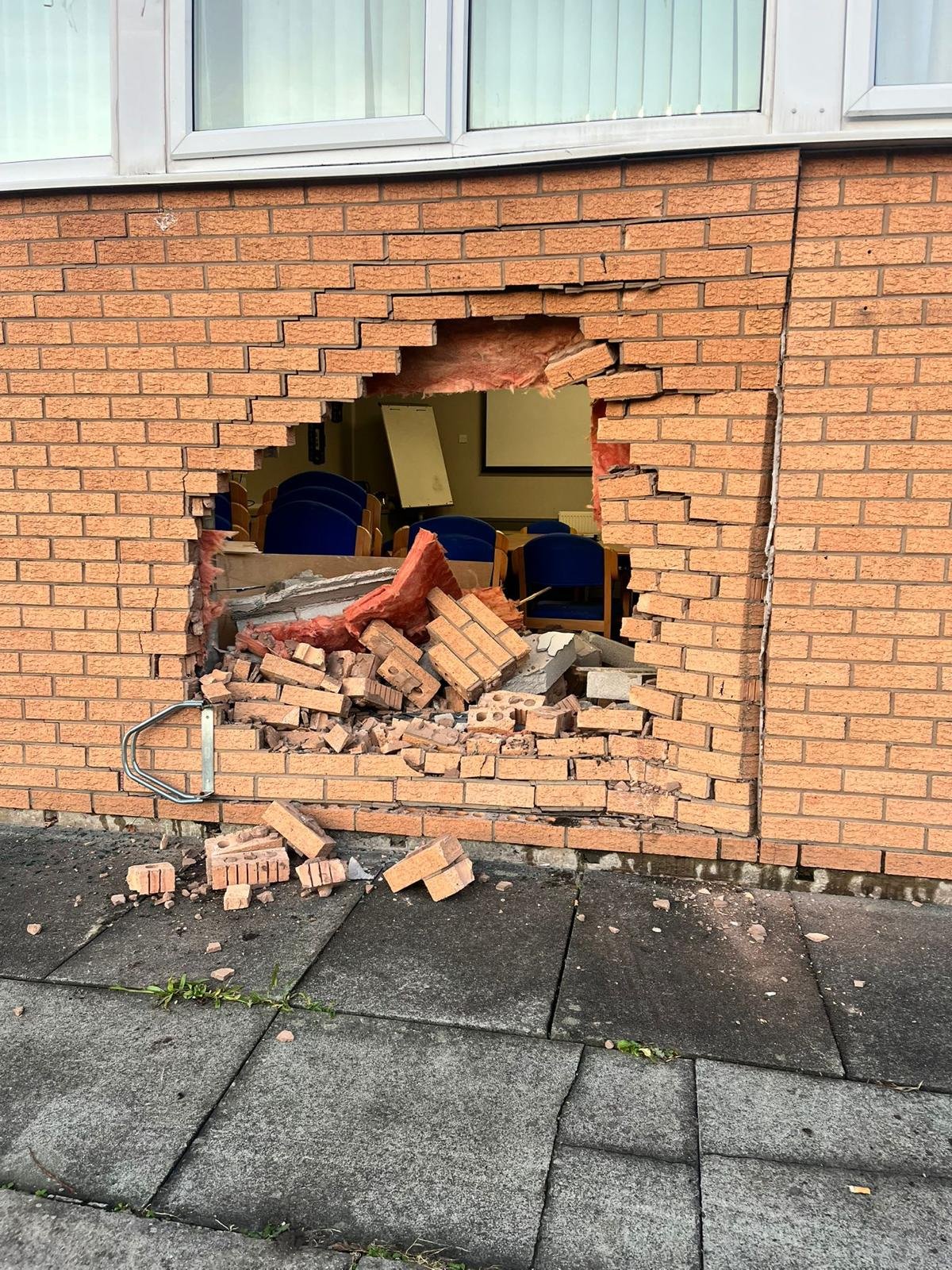 Hole through a brick cavity wall of a commercial building in Ellesmere Port after a vehicle impact, with bricks and insulation on the pavement