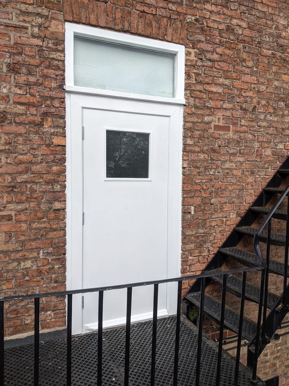 Completed FD30 timber fire exit door painted white, installed at the top of an external steel escape staircase on a brick commercial building in Chester