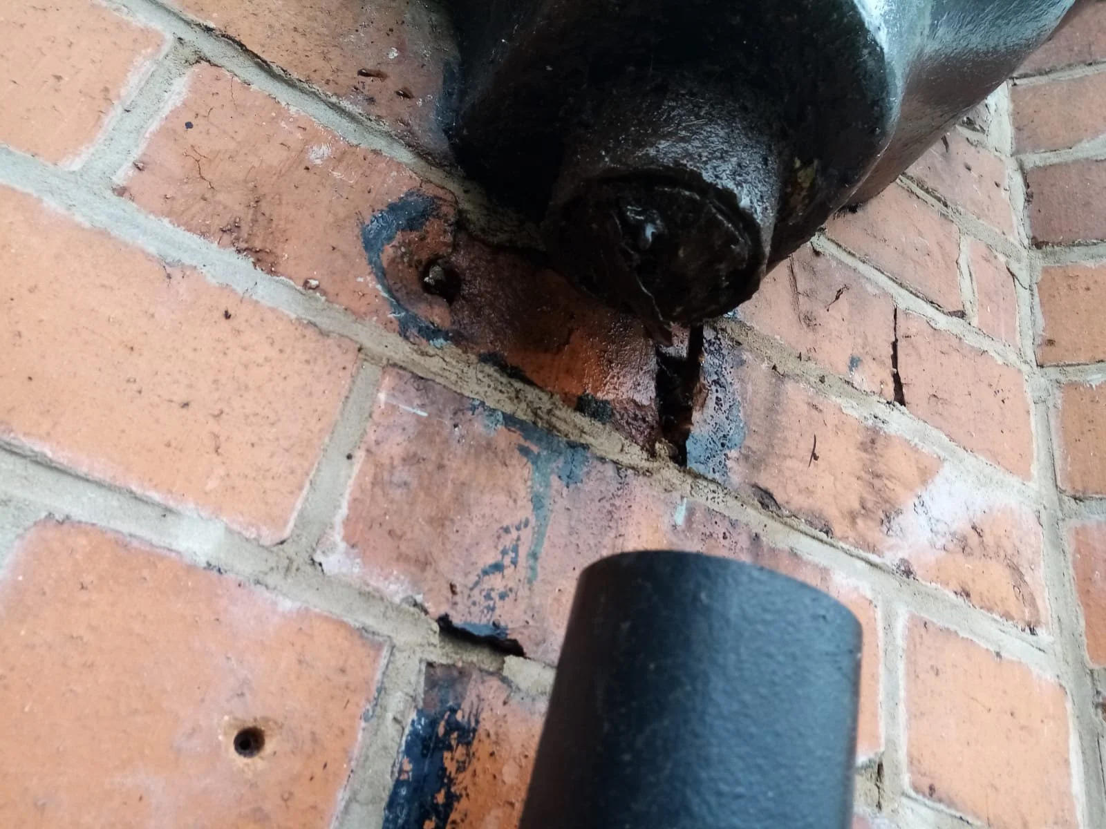 Outlet at the base of a cast-iron rainwater hopper with the downpipe removed, and dark water staining on the brickwork behind it