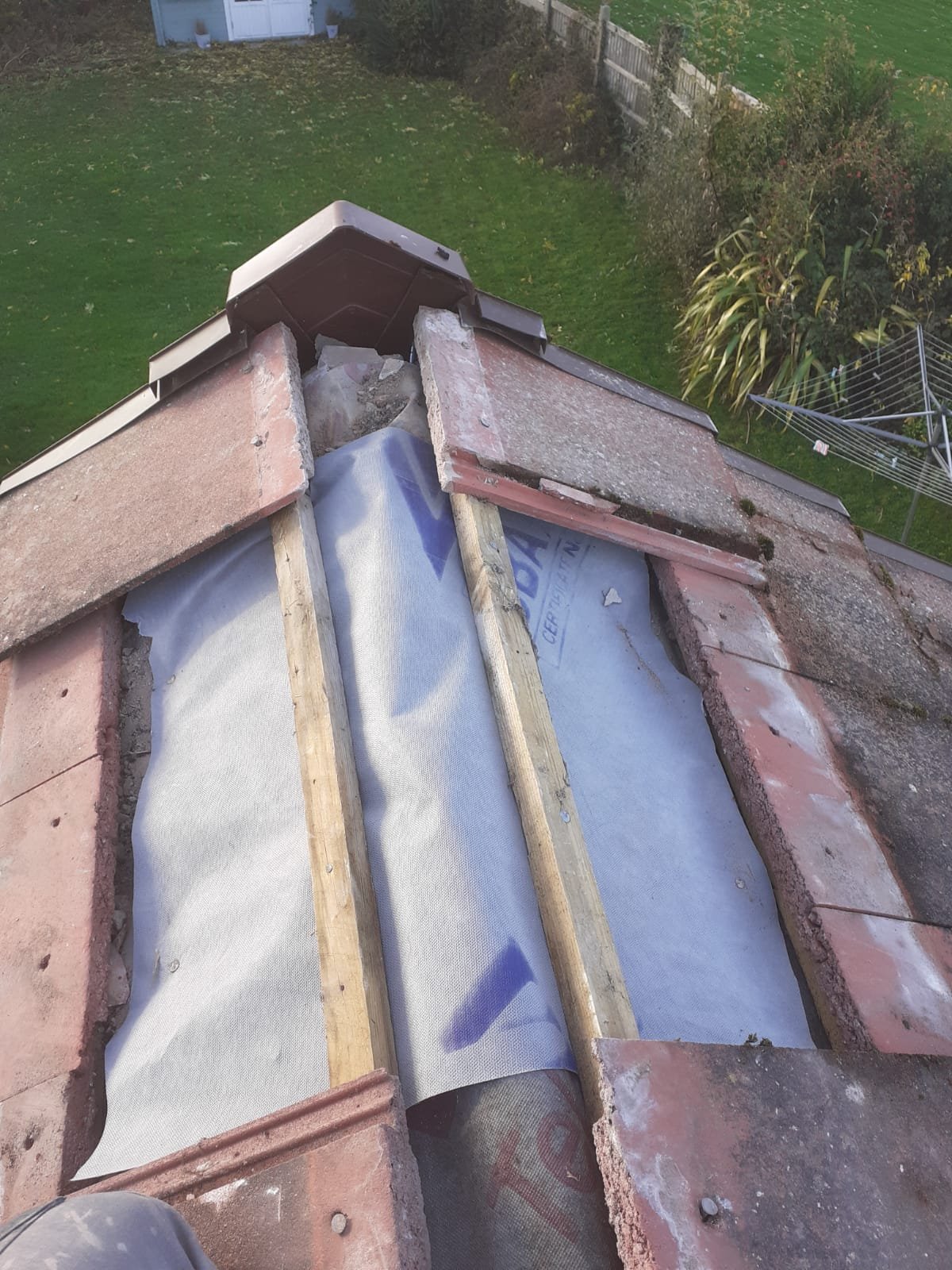Ridge of a tiled roof opened out with the ridge tiles lifted, showing broken-out mortar bedding, new ridge battens and the membrane beneath