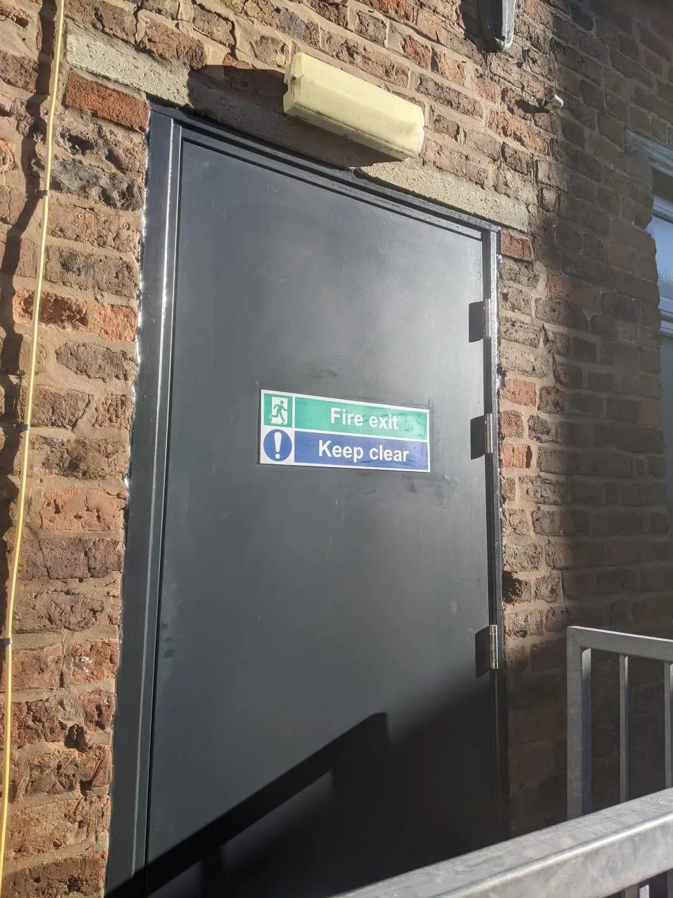 Completed black steel fire exit door installed in a brick opening at commercial premises in Birkenhead, with fire exit keep clear signage and an external bulkhead light above