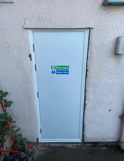 Completed white steel fire exit door installed in a rendered external wall at a care setting in Ellesmere Port, with fire exit keep clear signage