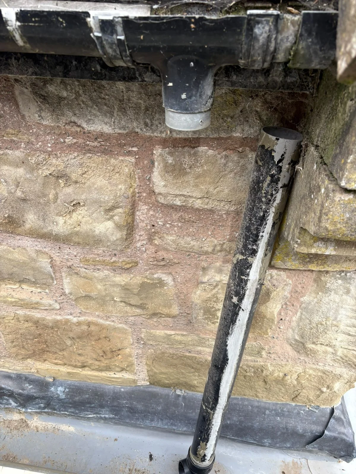 Storm-damaged downpipe detached from the gutter outlet and leaning against a coursed stone wall