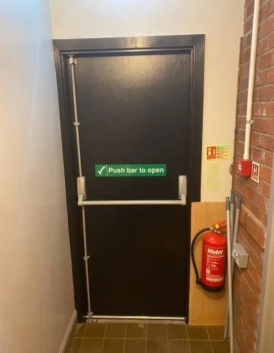 Completed FD30 timber fire exit door at an industrial unit in Buckley, decorated in black with push bar to open signage fitted