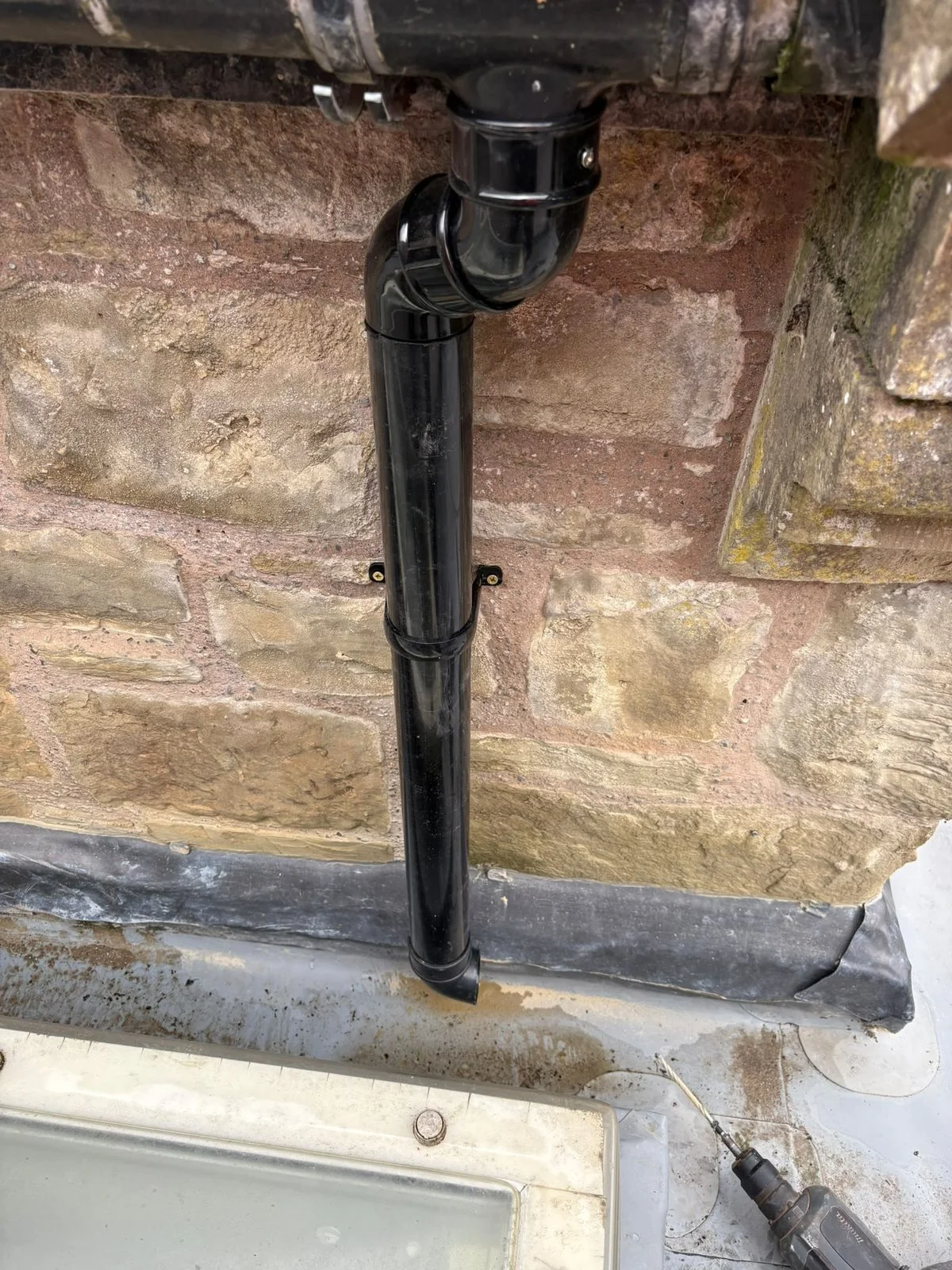 New black uPVC downpipe fitted to a coursed stone wall, offset from the gutter outlet above it