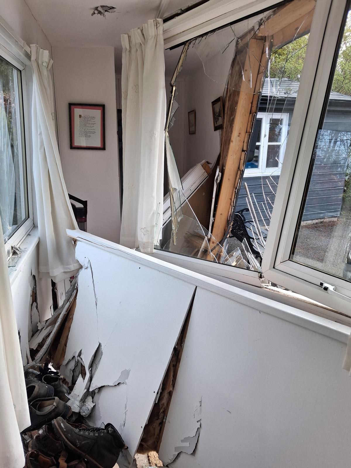 Inside a porch after a vehicle impact, with the wall lining split open, a broken window frame and glass and timber on the floor