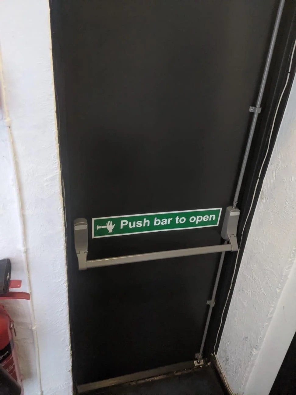 Interior view of a steel fire exit door showing the three-point panic bolt with top and bottom shoot bolts and push bar to open signage