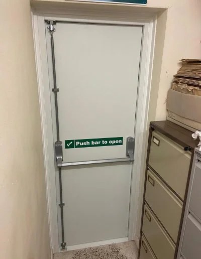 Interior view of a white steel fire exit door showing the three-point panic bolt with top and bottom shoot bolts and push bar to open signage