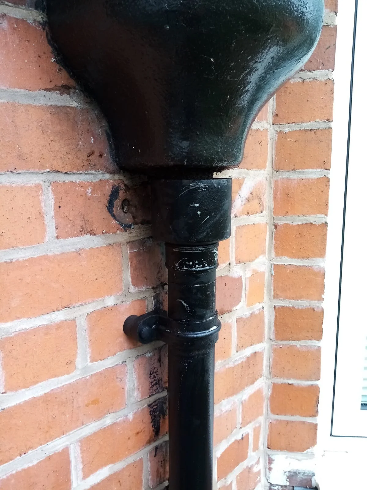 Cast-iron downpipe refitted beneath a rainwater hopper on a red brick wall, held by a pipe bracket