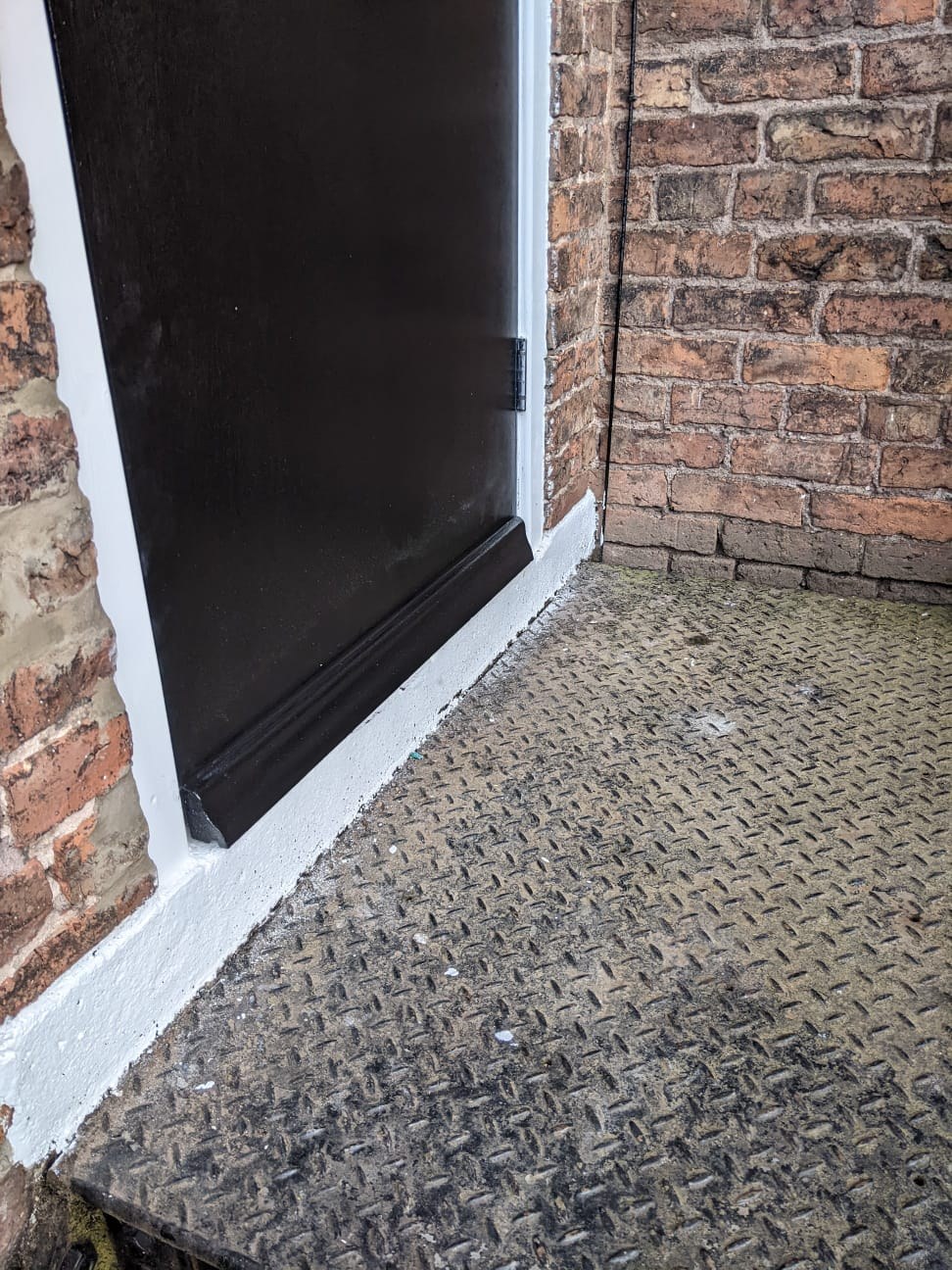 Close-up of a fire exit door threshold showing the fitted weather bar at the foot of the door leaf and the sealed junction with the external steel landing