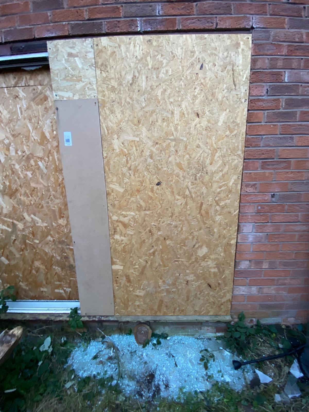 Patio door of a brick house boarded up with OSB sheets after a break-in, with broken glass on the ground outside