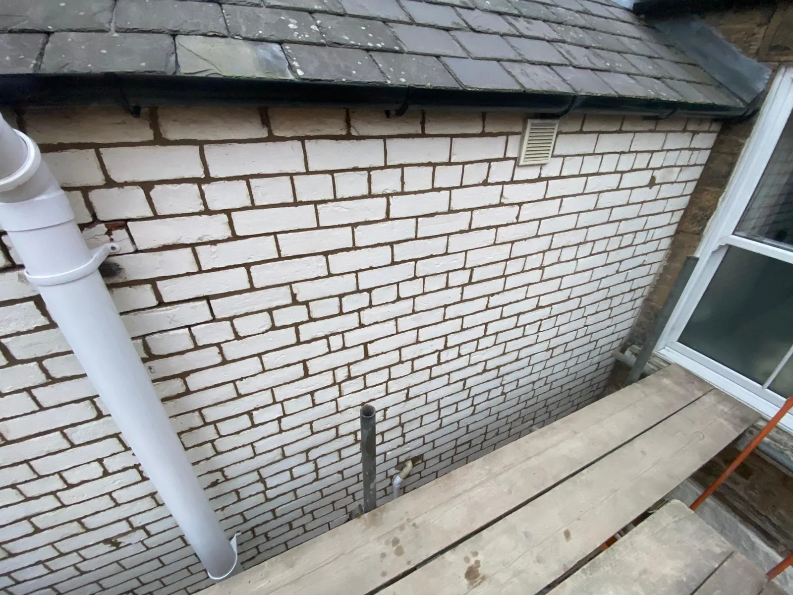 Painted brick wall under a slate roof, seen from the scaffold, with cracked brickwork beside the downpipe