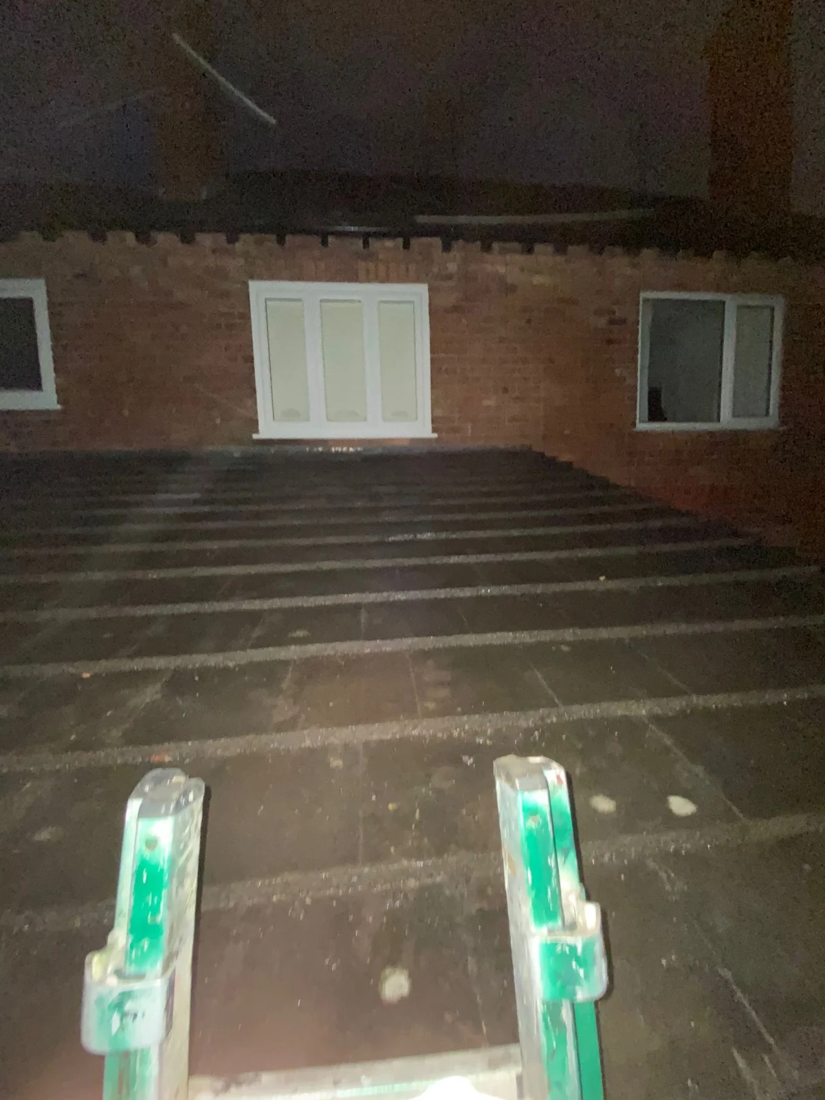 Flat roof at night, seen from the top of a ladder, with a brick wall and windows beyond