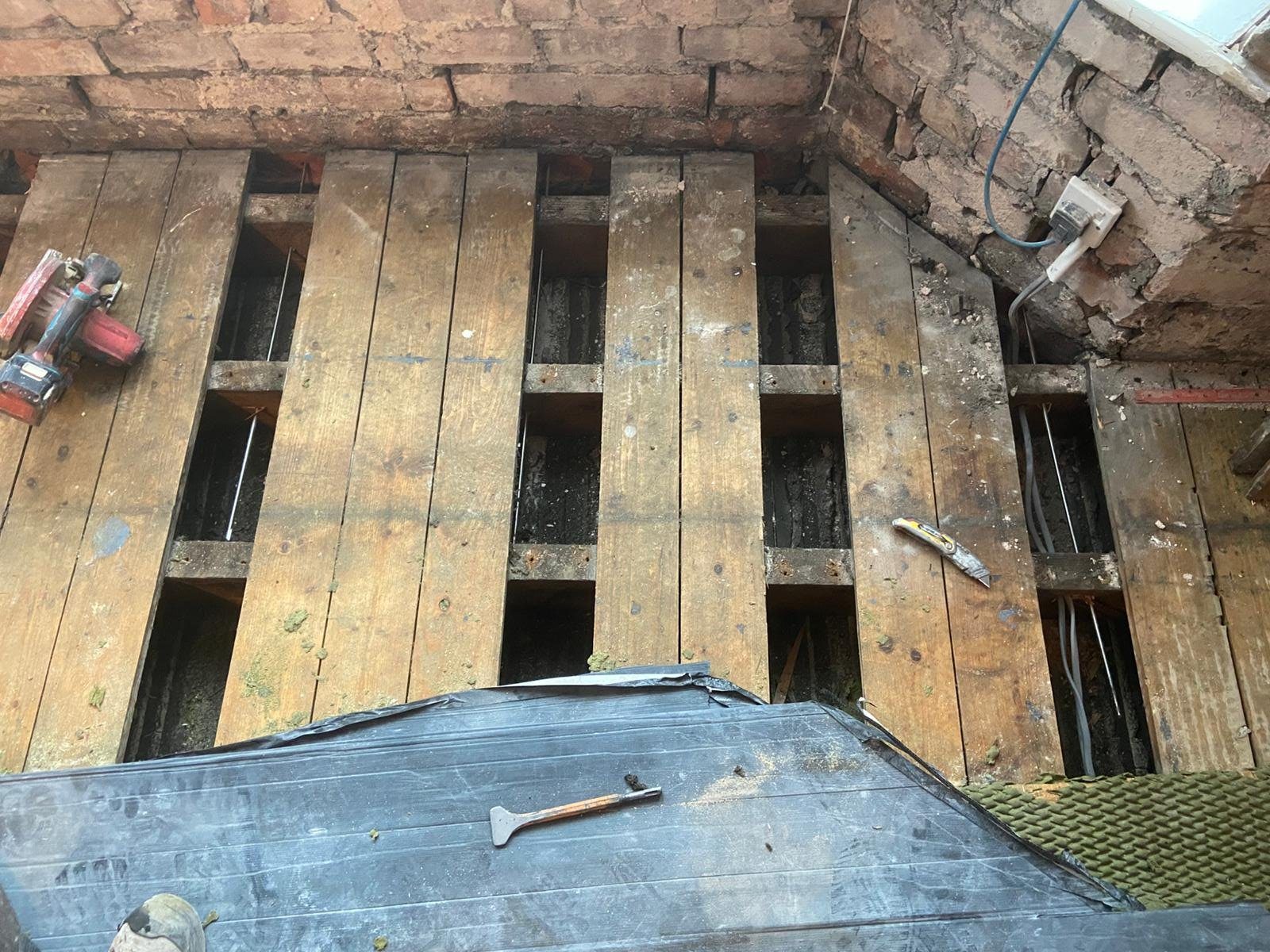 Floorboards lifted in strips beside the bay wall, showing steel bow ties fixed between the floor joists