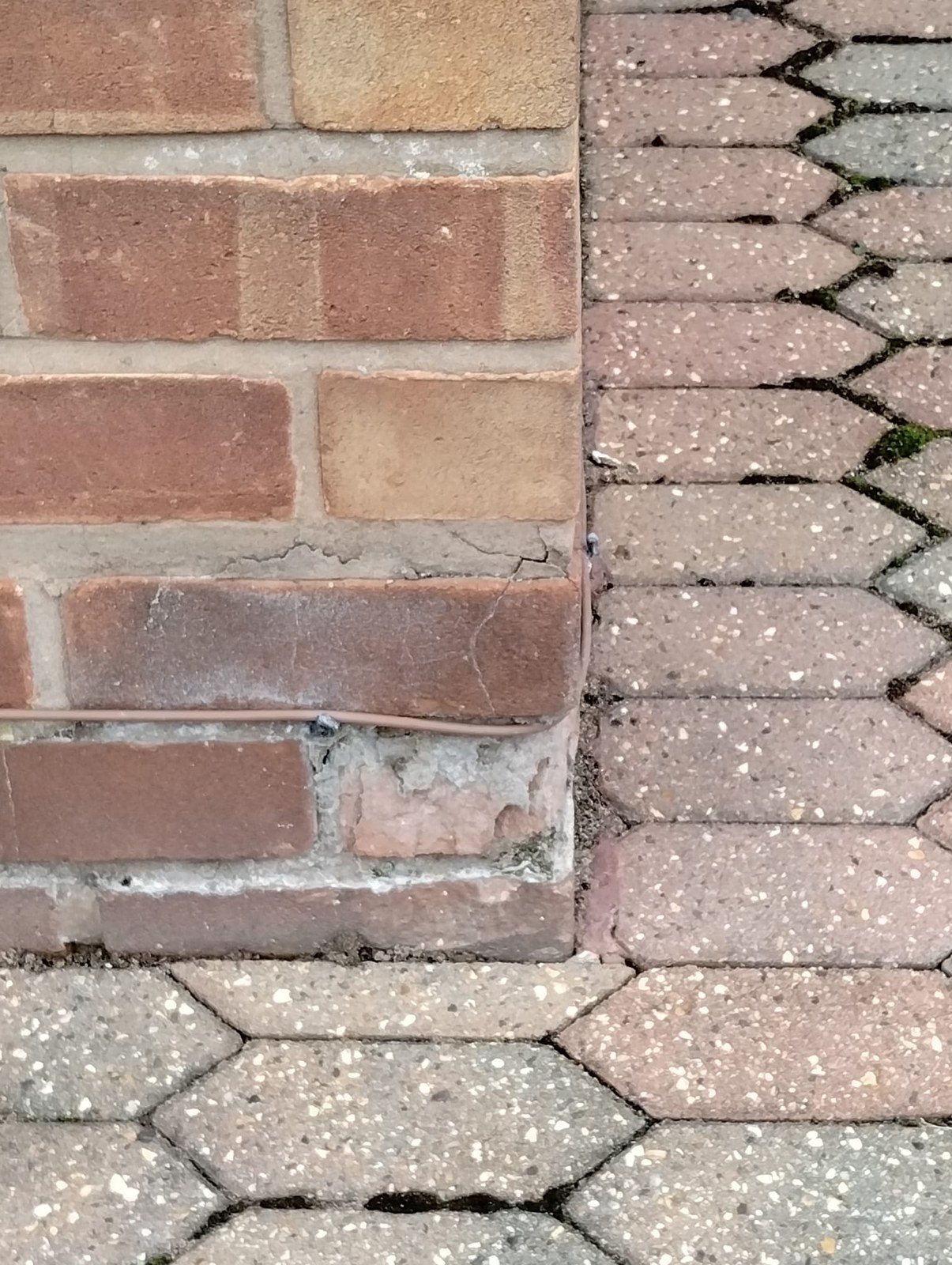 Brick repair in Flintshire – cracked joint and broken brick face before Red brick corner at ground level beside block paving, with a cracked mortar joint, a crack across one brick and a broken brick face below it