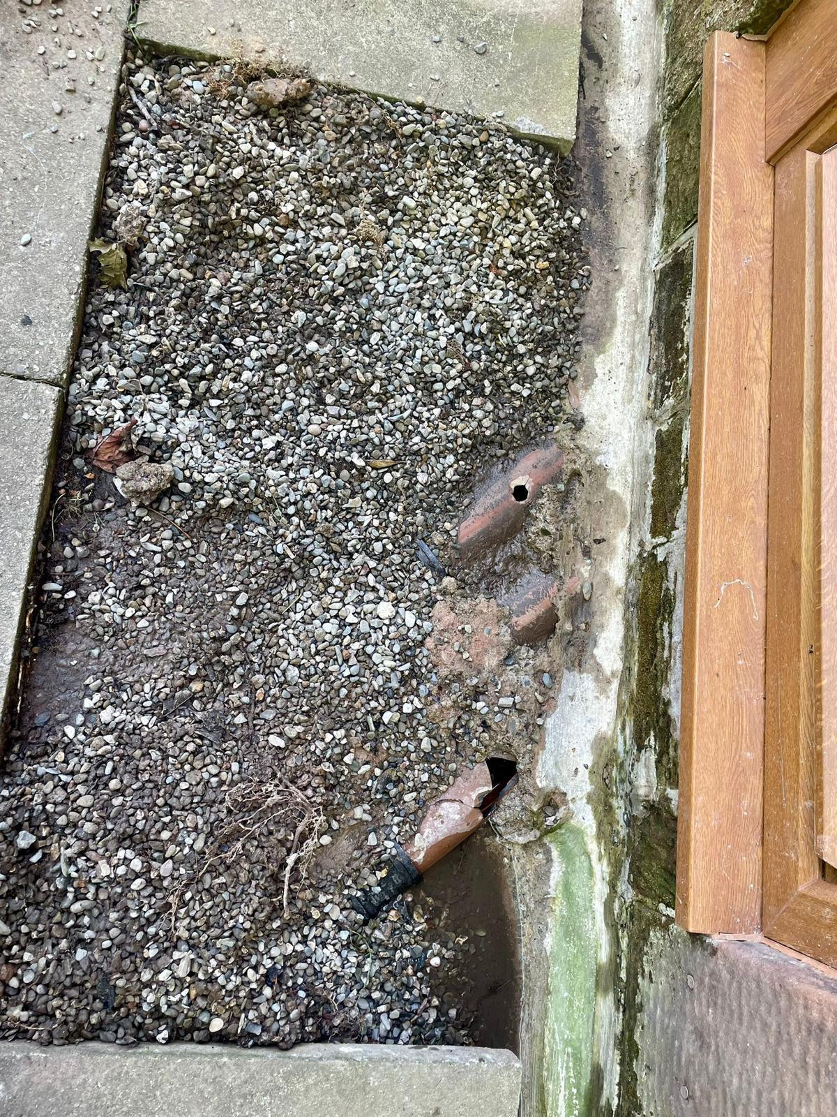 Excavation alongside a front doorstep exposing an old clay drain run against the wall of a stone-built house
