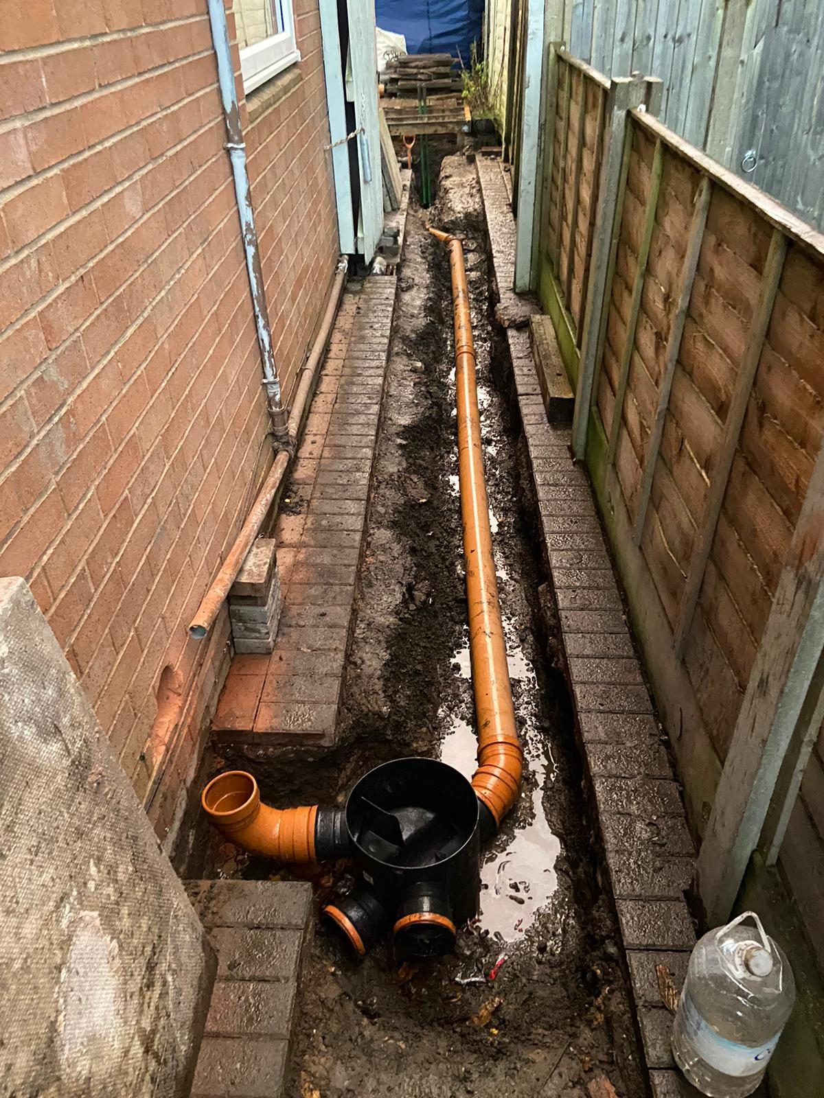 Trench along a narrow side passage beside a brick wall, with a new orange drainage pipe laid its full length and a new black inspection chamber in the foreground