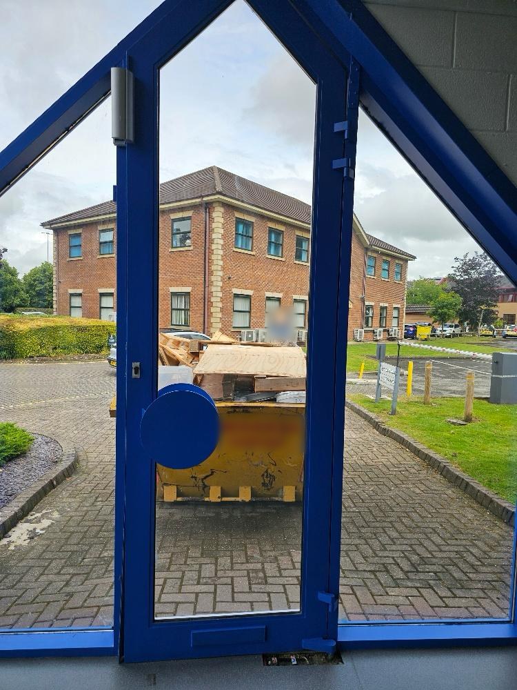 View from inside an office entrance through a tall blue-framed glazed door with a round pull plate, towards a car park and office buildings