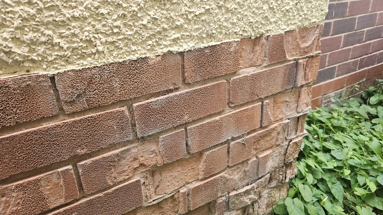 Corner of a brick plinth below a roughcast rendered wall, with the faces of several bricks blown off and the orange inside of the bricks exposed