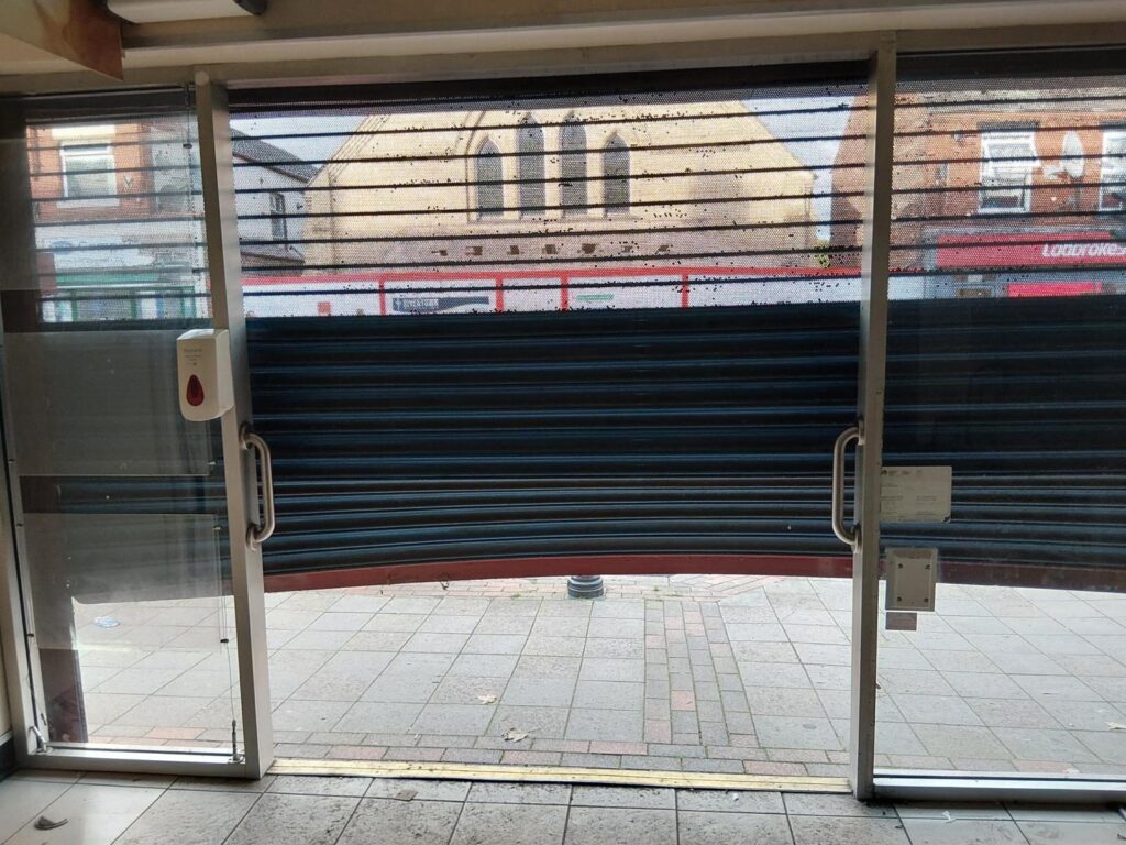 View from inside a shop entrance after a break-in, with the roller shutter's bottom edge bent up off the pavement