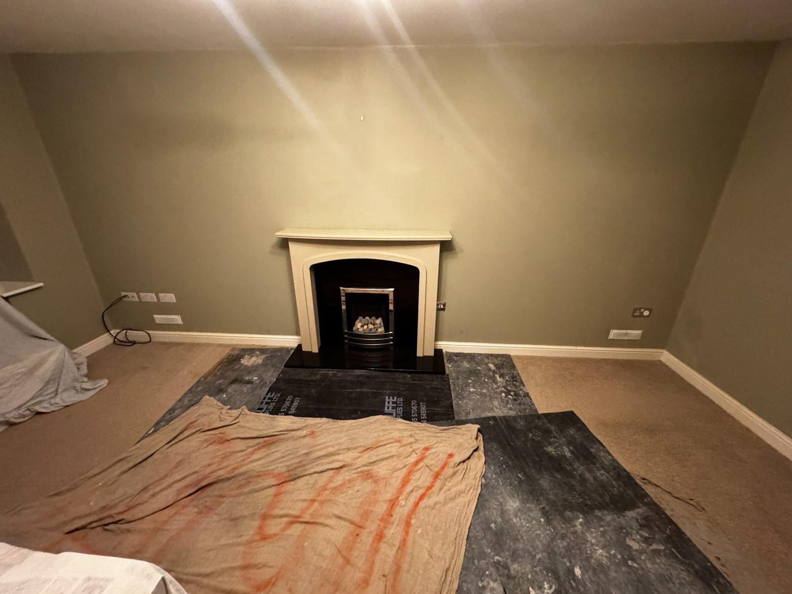 Cream stone fireplace surround with an arched opening, chrome inset fire and raised black hearth in a flat painted wall, with floor protection boards and a dust sheet on the carpet