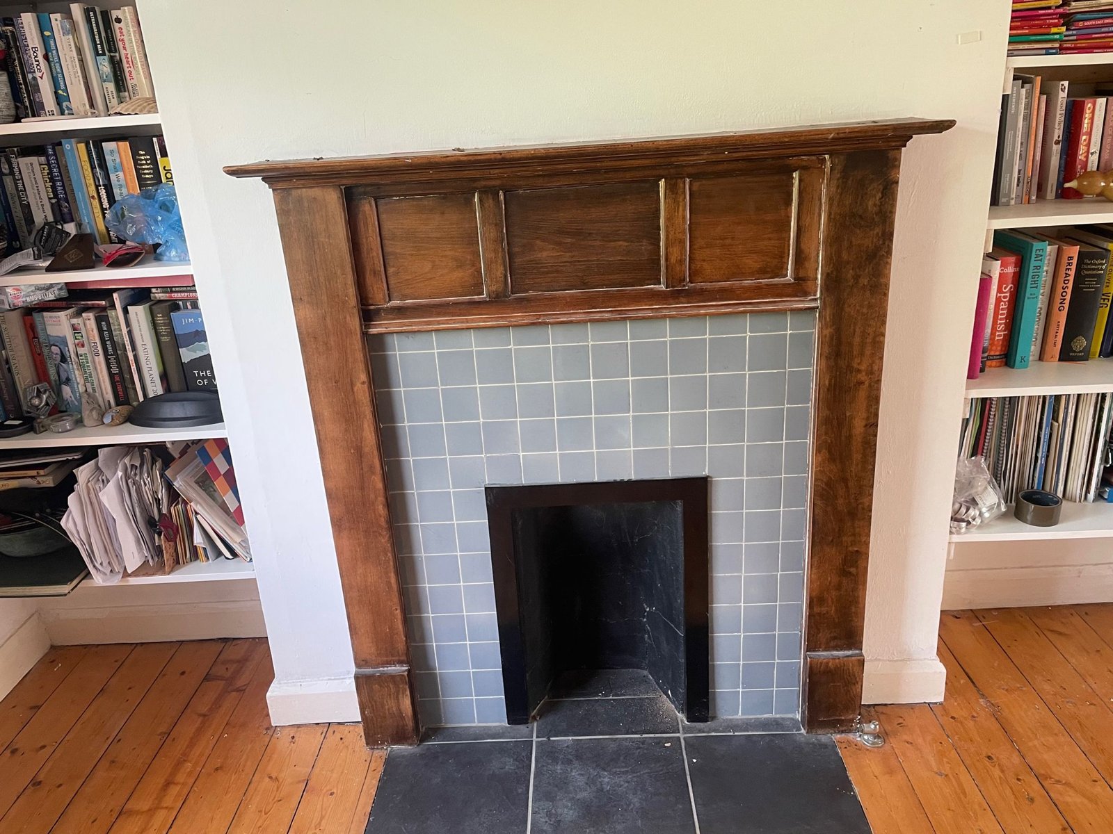 Dark timber fireplace surround with blue-grey tiles, a small black opening and a slate hearth, between fitted bookshelves on a pine floor