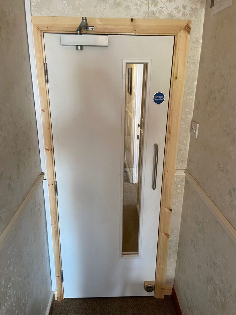 White fire door with overhead closer, vision panel and new timber architraves in a converted house hallway