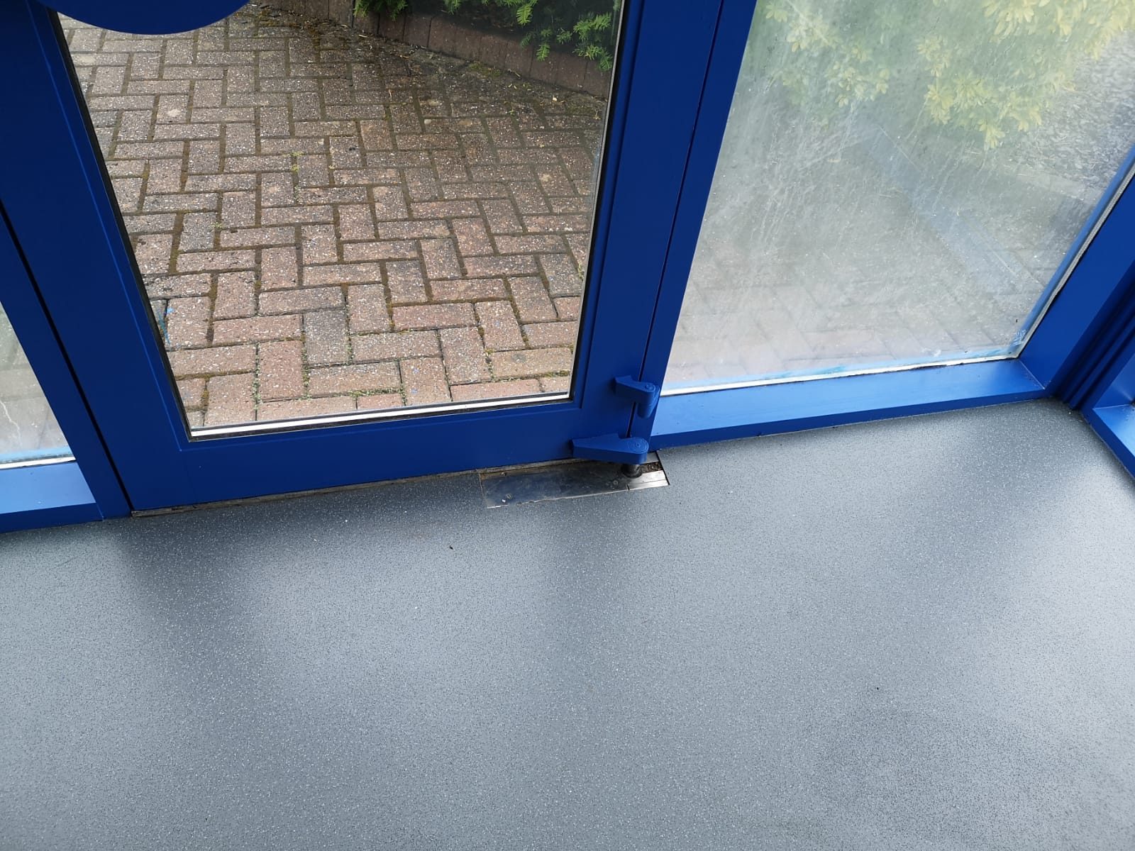 Foot of a blue-framed glazed door from inside, shut onto a grey floor, with its hinges and a metal plate in the floor at the hinge side