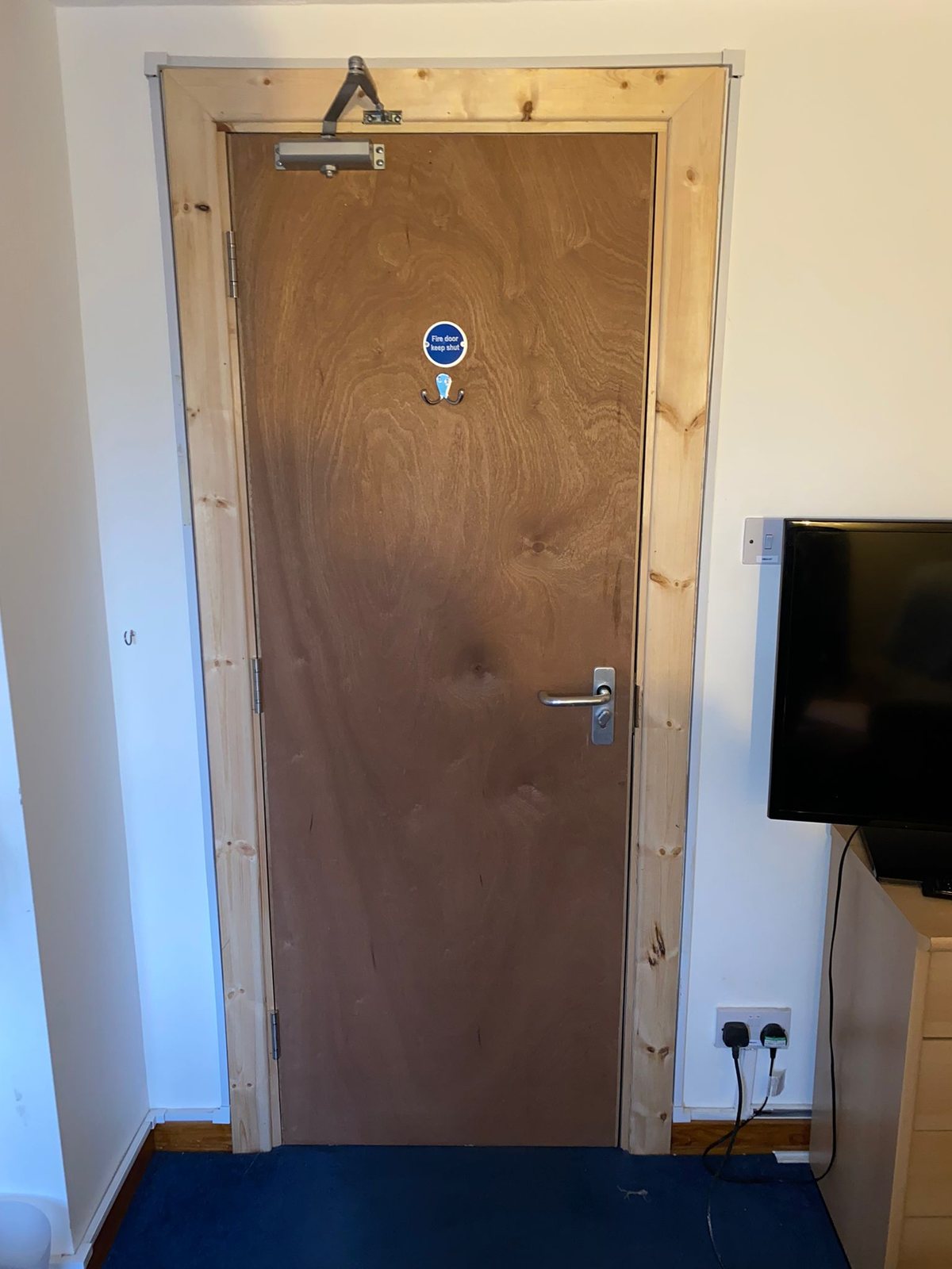 Plywood flush FD30 bedroom fire door seen from inside the room, with an overhead door closer, a lever handle with thumb-turn and a fire door keep shut sign, in an undecorated timber frame