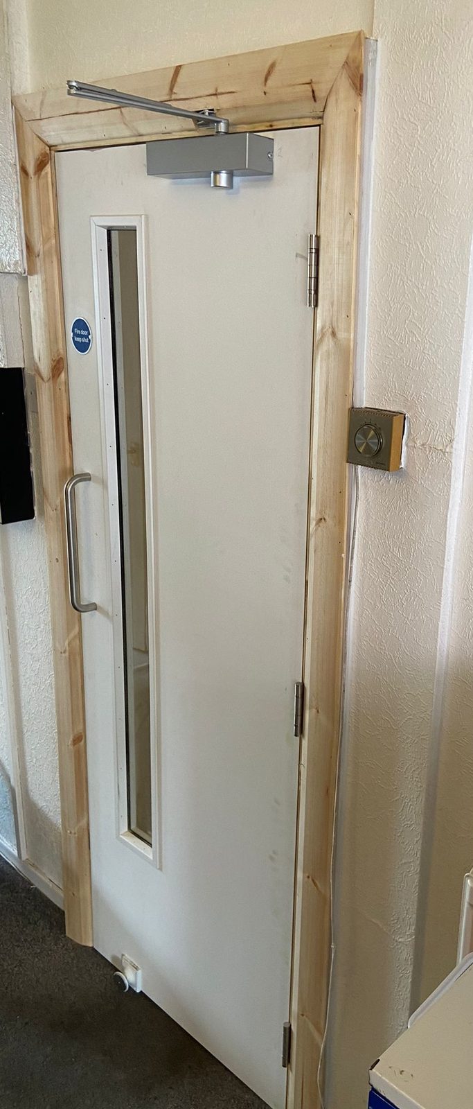 White FD30 corridor fire door with a vision panel, overhead door closer and pull handle, and a hold-open device at the foot of the leaf
