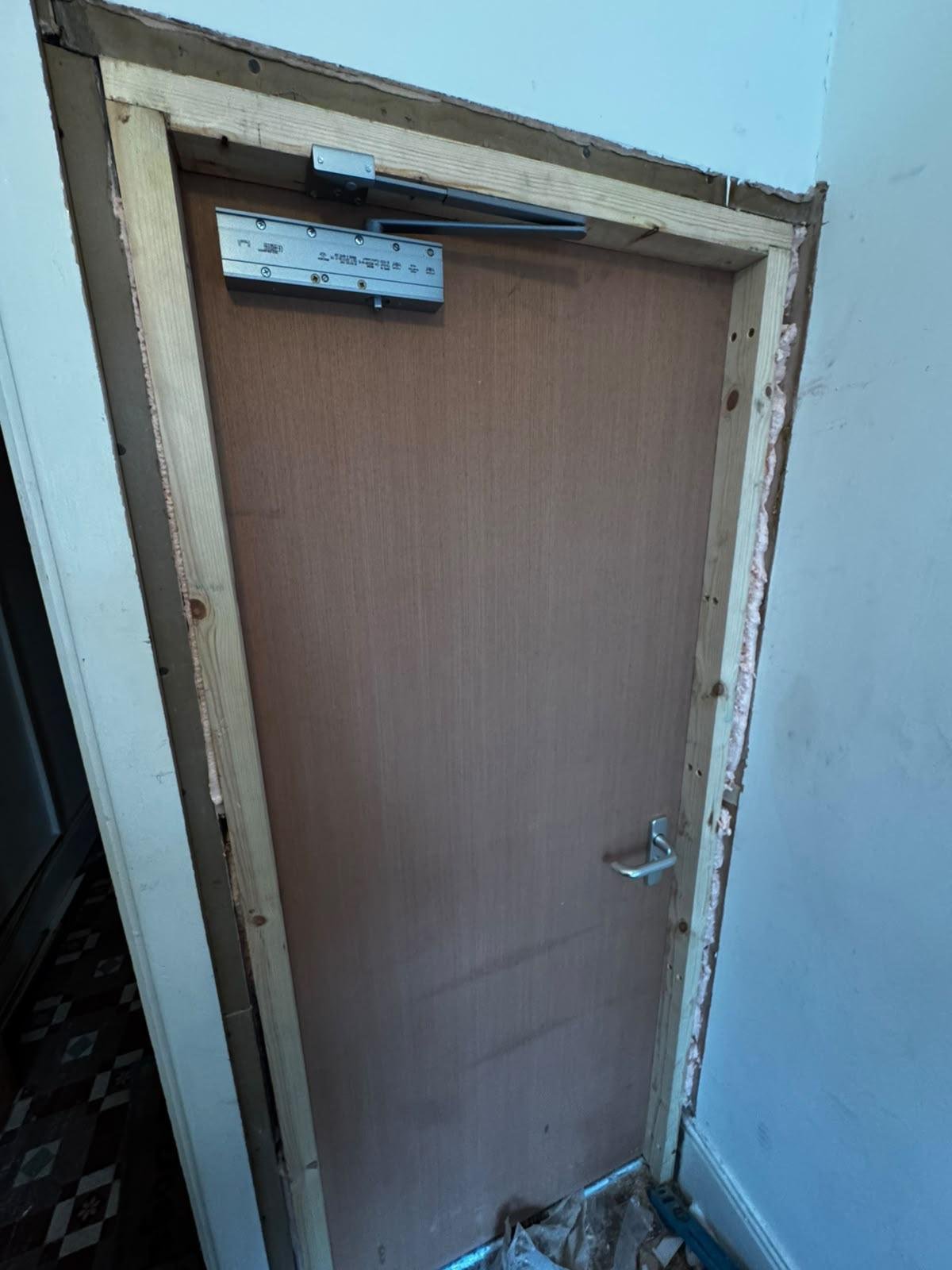 New plywood flush FD30 fire door and timber frame fitted in an existing opening, with an overhead door closer and lever handle, before the architraves are fitted