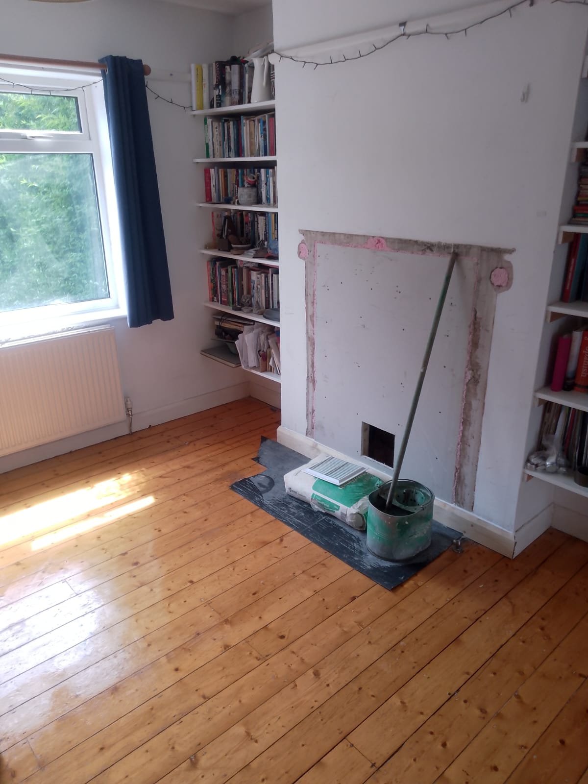 Chimney breast with the fireplace removed and the opening boarded over, a square vent opening cut near the floor, and a plastering bucket on floor protection