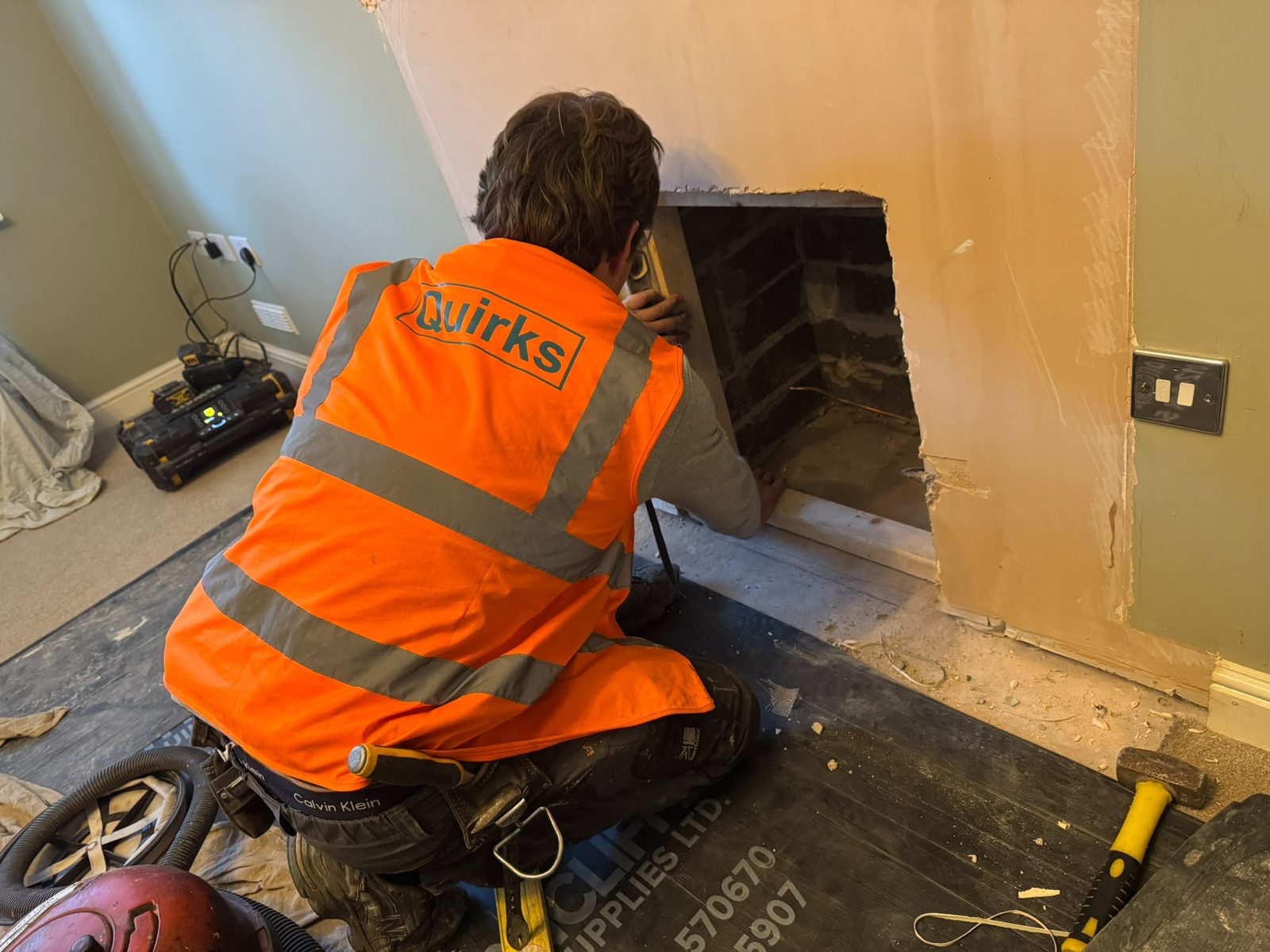 Quirks operative in a high-visibility vest using a spirit level on the timber frame in a fireplace opening, with the surround and hearth removed and floor protection down