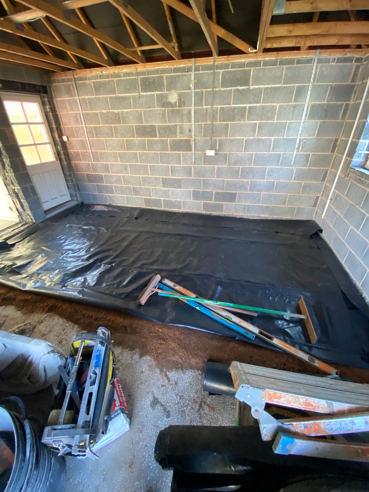 Damp-proof membrane laid over a sand blinding across a garage floor slab, with bare blockwork walls and exposed roof trusses