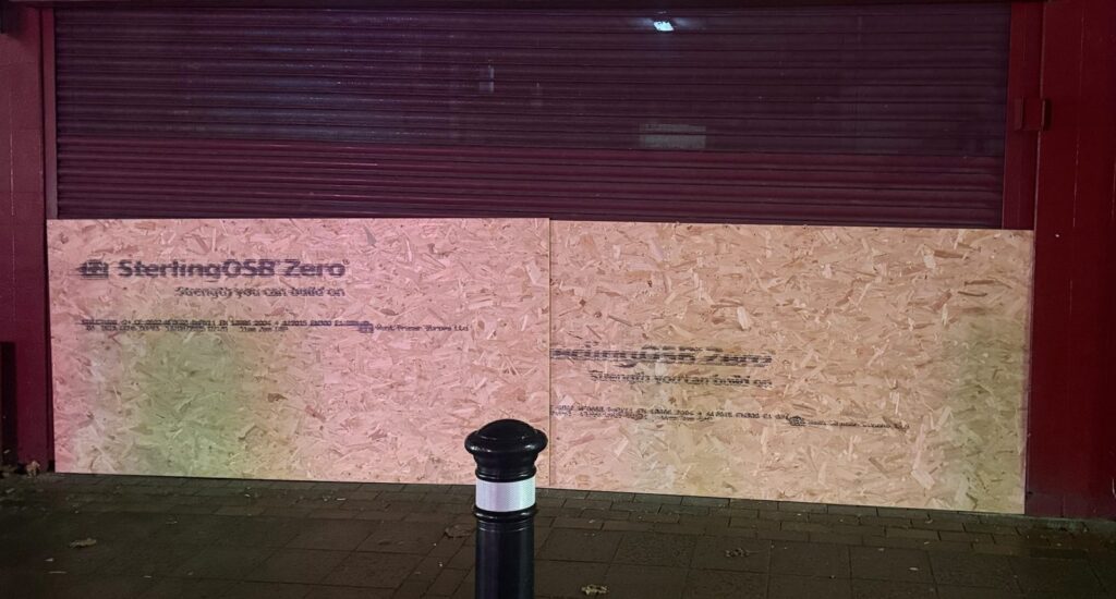 Shop front in Flintshire secured with OSB boards fixed across the lower half below a closed roller shutter