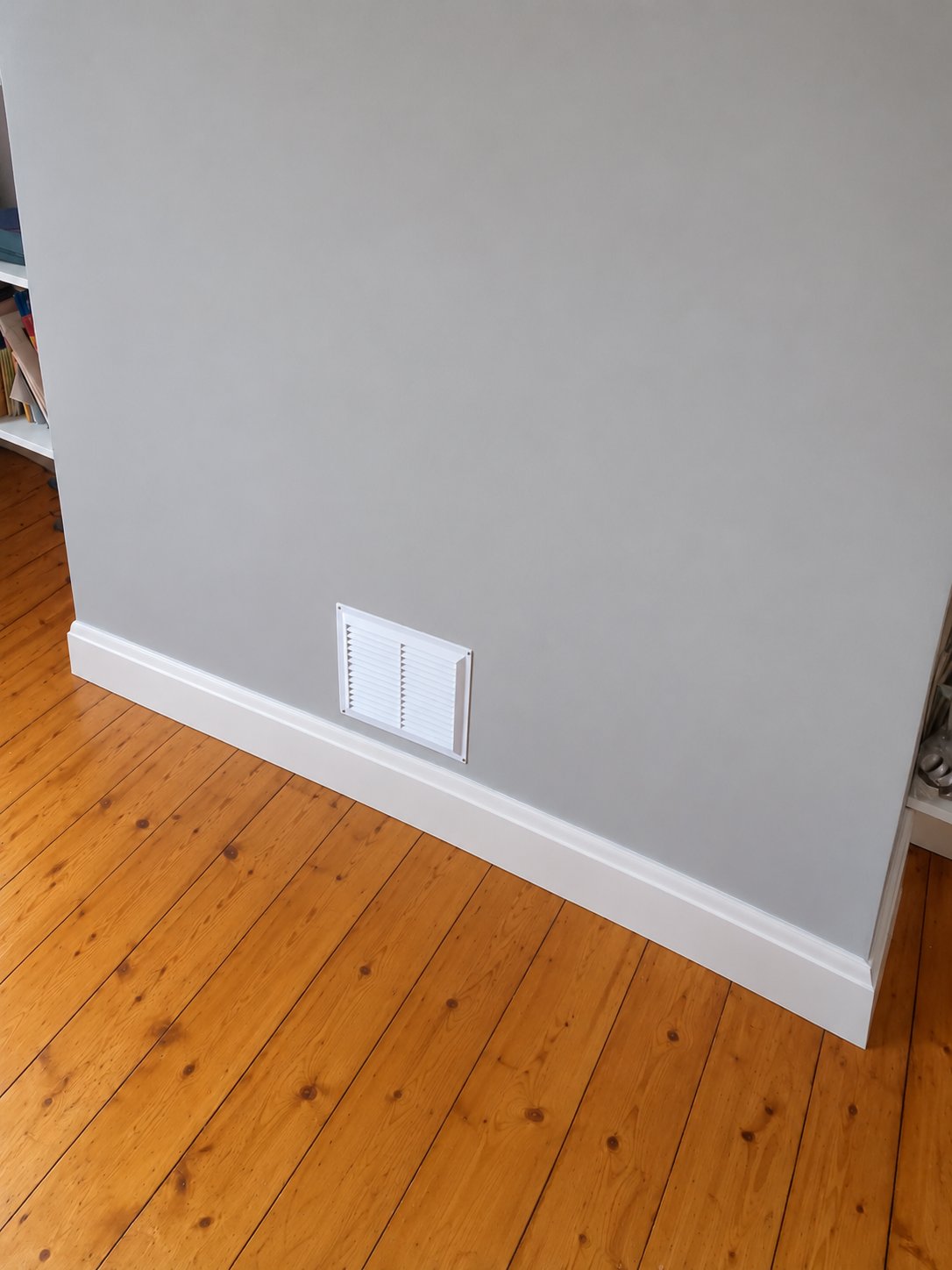 Grey painted chimney breast with a white ventilation grille above new white skirting board, on a pine floor