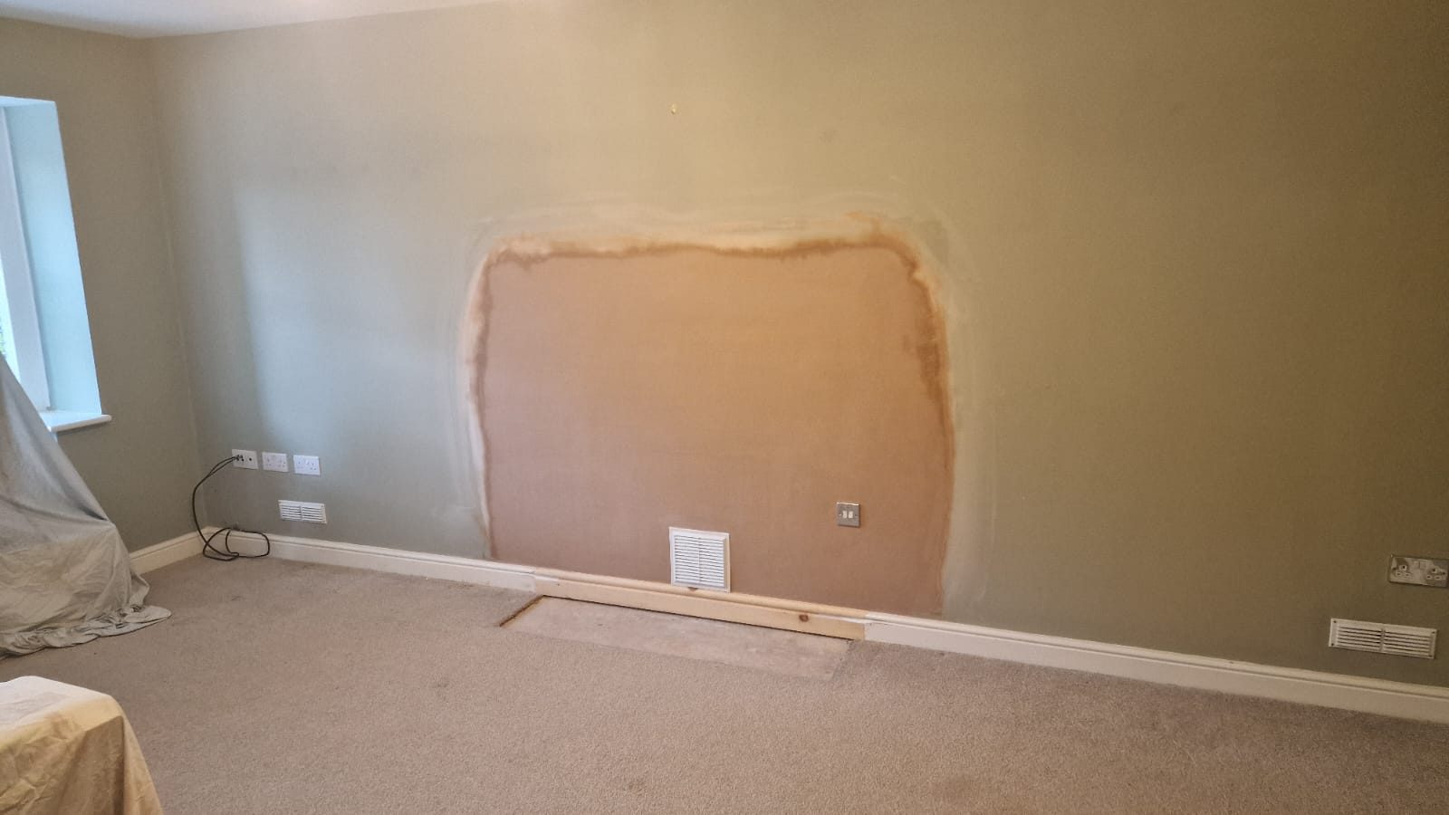 Freshly skimmed section of wall where a fireplace was removed, with a white ventilation grille above new timber skirting and a bare strip of floor in front