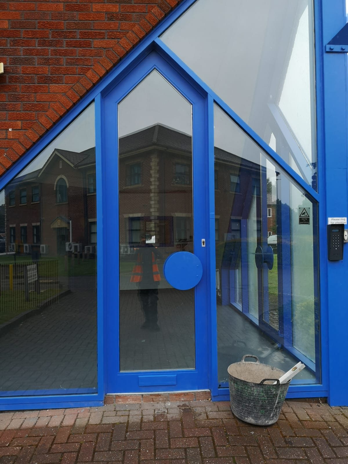 Tall blue-framed glazed entrance door shut, seen from outside, with a keypad entry panel beside it and a builder’s tub on the paving