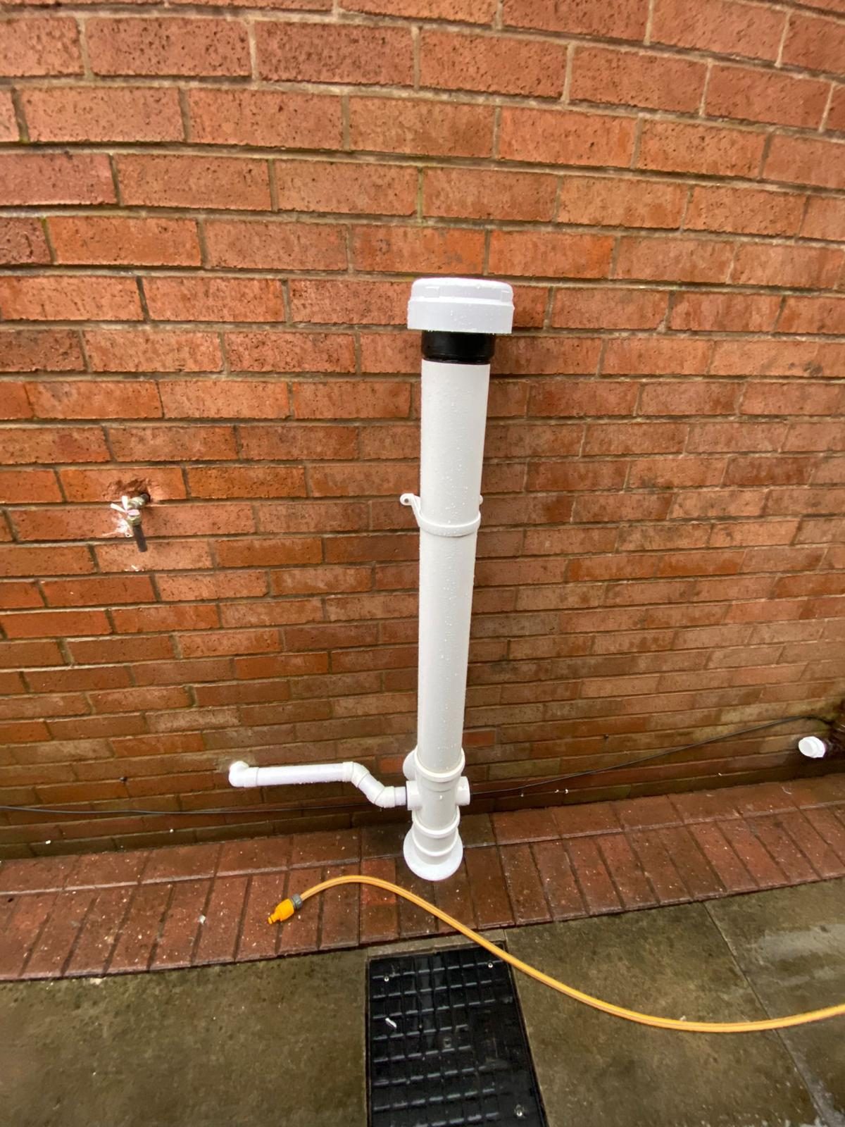 Short white external drainage stack on a brick wall, capped with an air admittance valve, with a small waste pipe coming out of the wall into its base