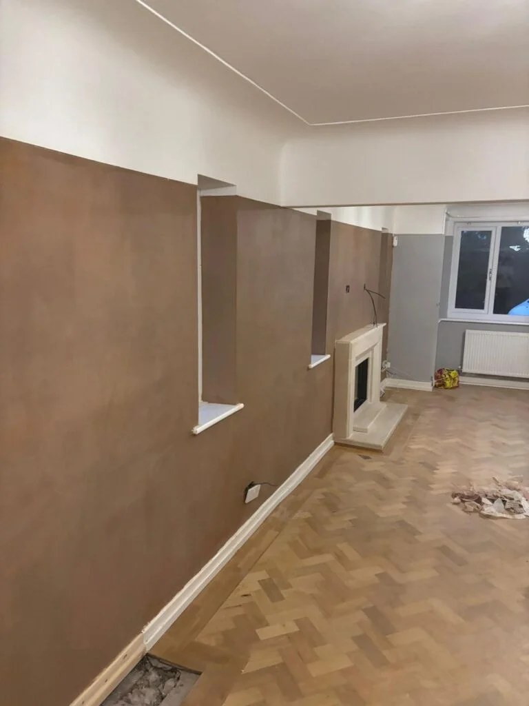 Long wall in fresh brown plaster with two small window reveals, a cream stone fireplace further along and a gap in the parquet floor in the foreground