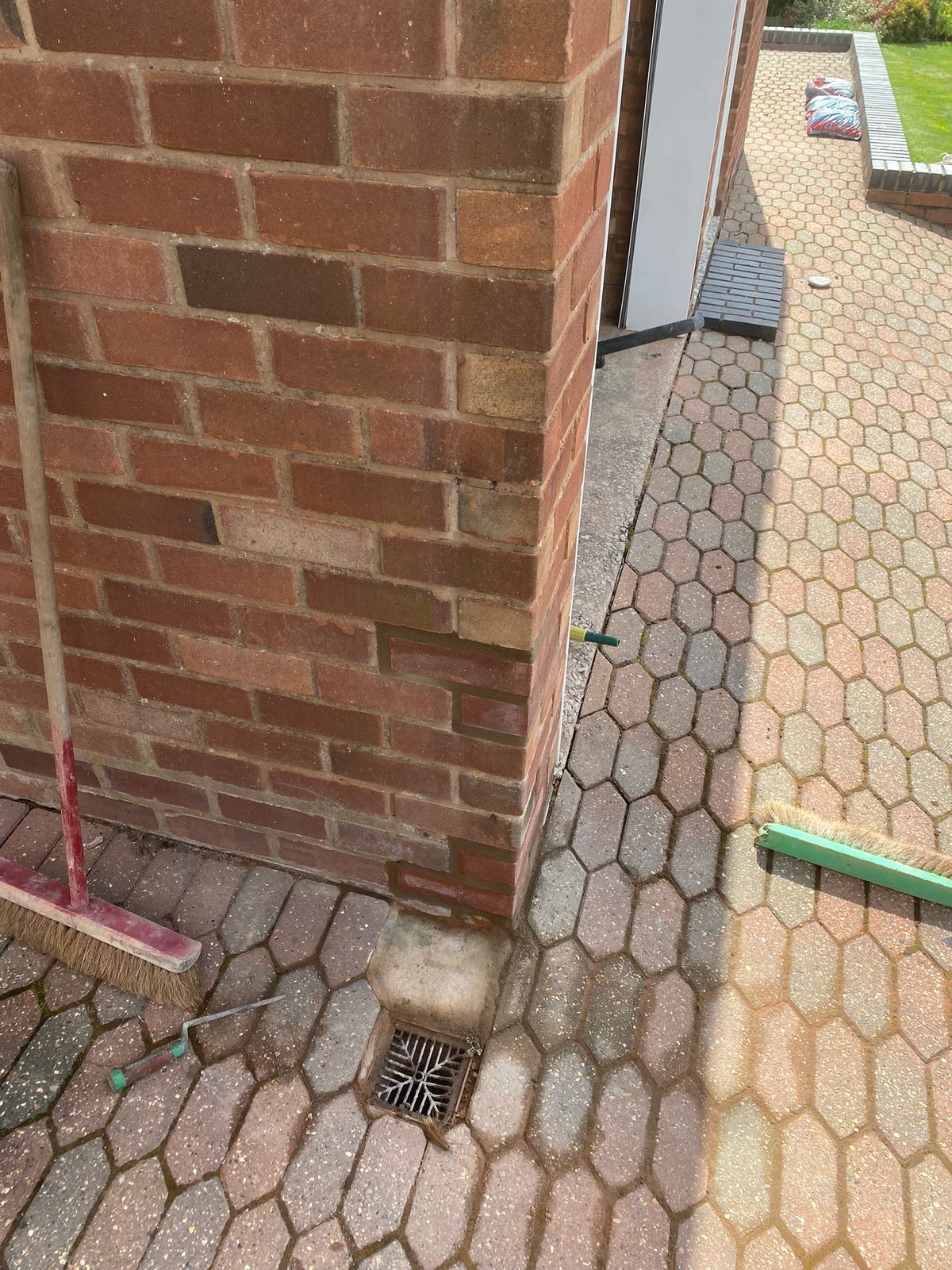 Brick repair in Flintshire – corner with replacement bricks after Brick corner above block paving with newer bricks set in at the base and higher up, a drain gully, a brush and a trowel on the paving