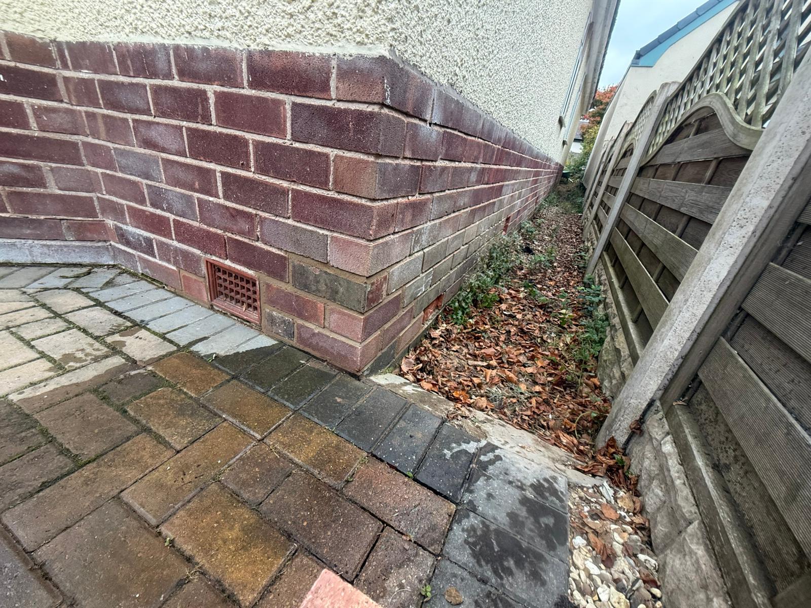 Corner of a brick plinth with new bricks in the lower courses, beside block paving, with the plinth continuing down a side passage