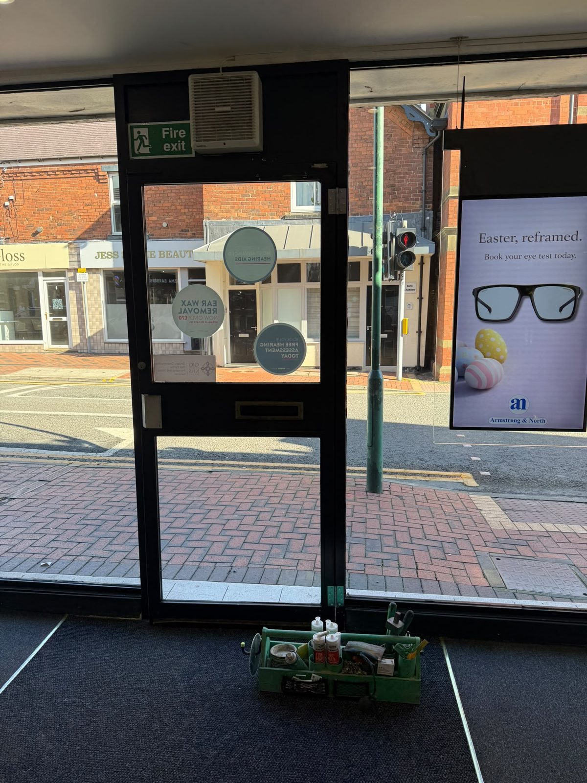 Shop entrance door re-hung on surface-mounted hinges after a temporary emergency repair, viewed from inside the shop