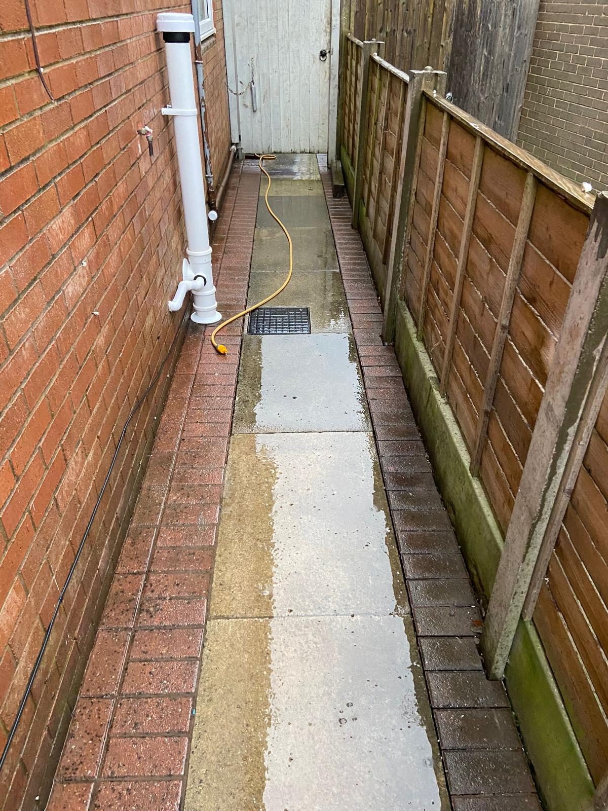 Narrow side passage with the paving slabs relaid between brick paviour edging, a drainage chamber cover set into the path and a white stack on the wall