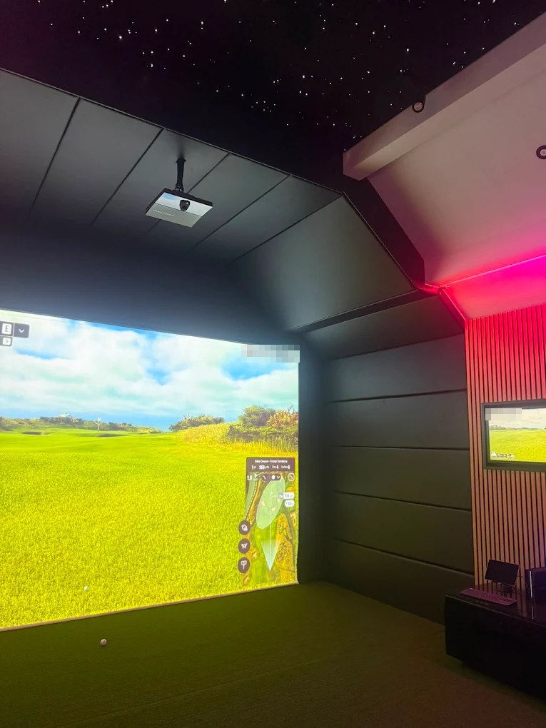 Golf simulator in a converted garage with a ceiling-mounted projector, full-width projection screen showing a golf course, acoustic panelled walls and a fibre-optic star ceiling