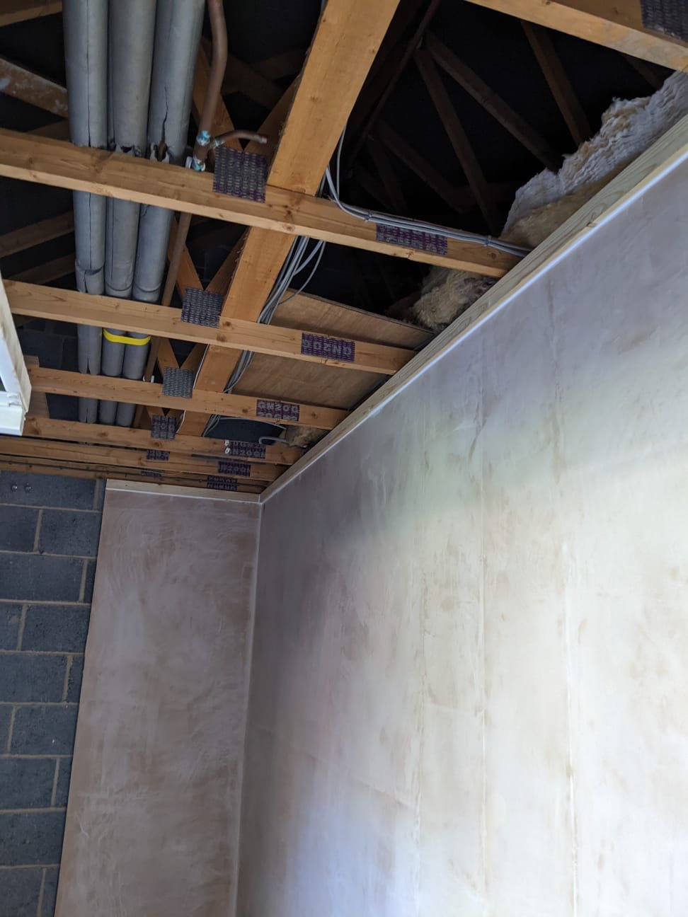 Freshly plastered walls and ceiling in a converted garage, with pipework and roof trusses visible above the finished ceiling line