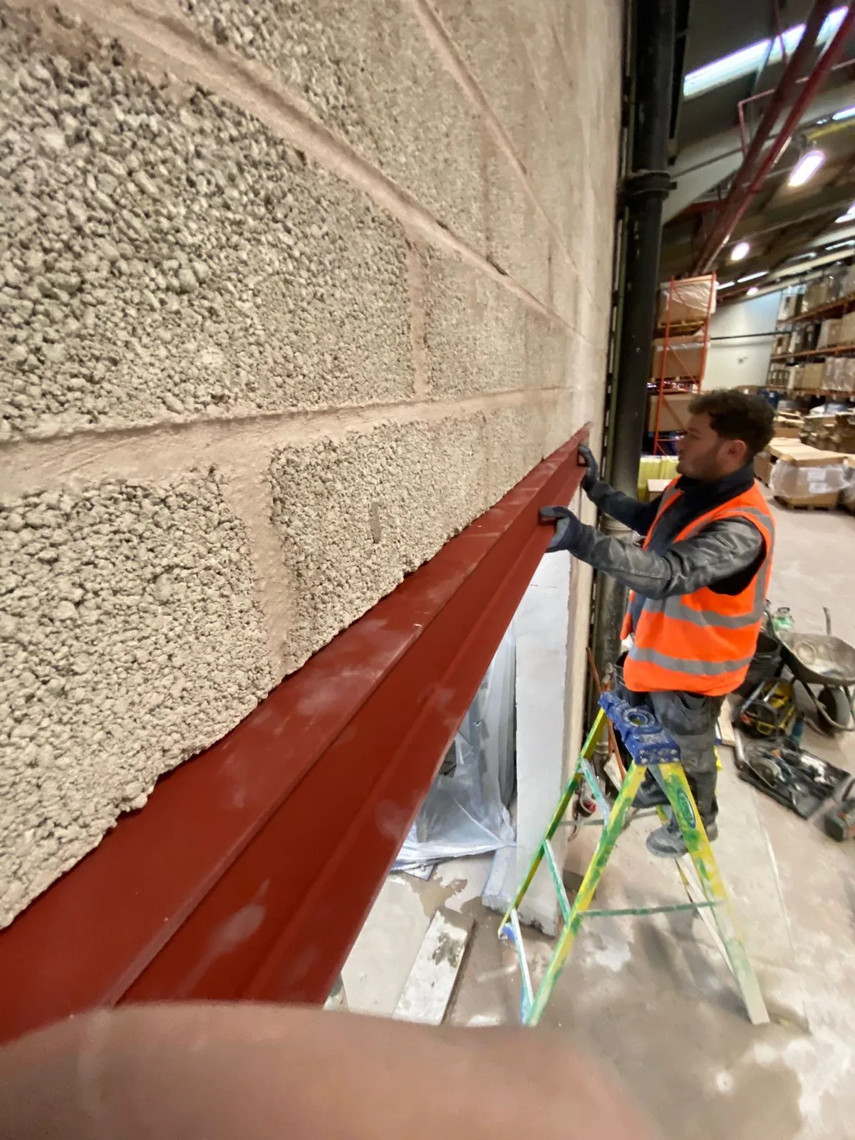 Operative on a step ladder positioning a red-oxide steel beam against a blockwork wall inside a working warehouse