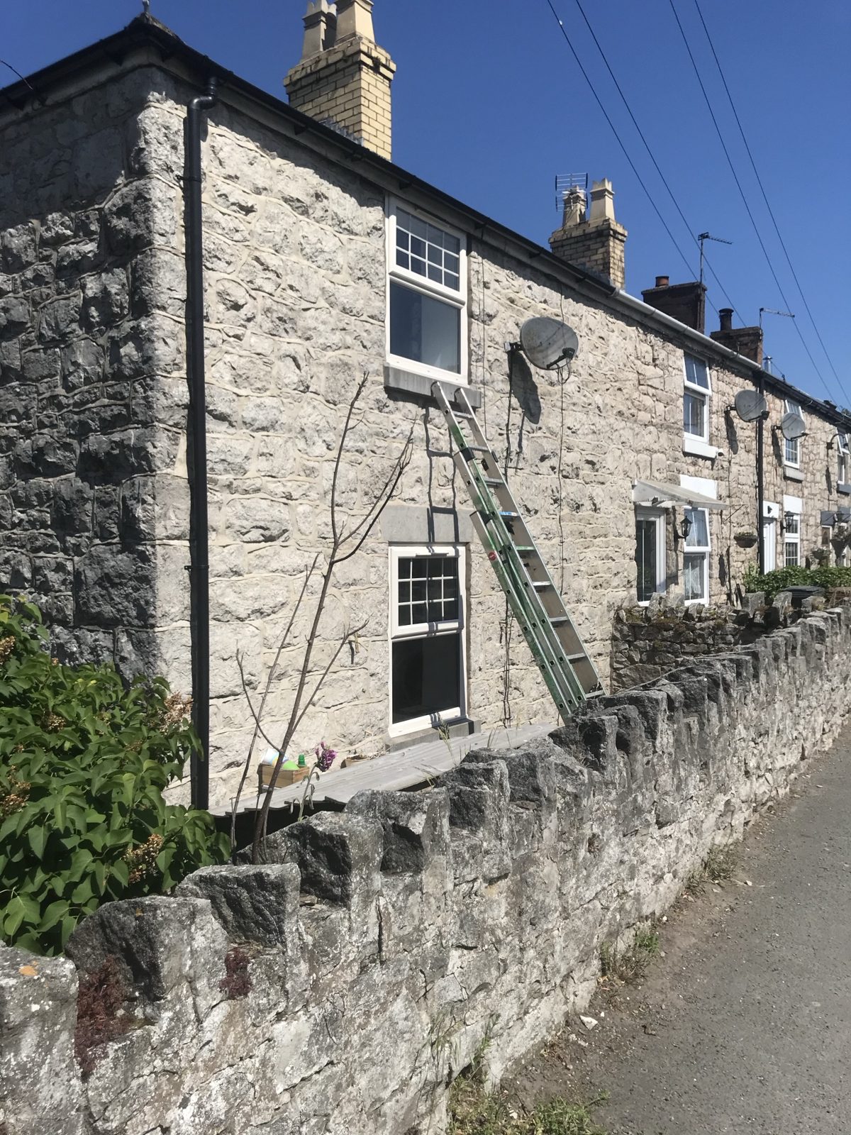 Row of stone-built terraced cottages with pale brick chimneys, white sash-style windows and a stone wall along the lane
