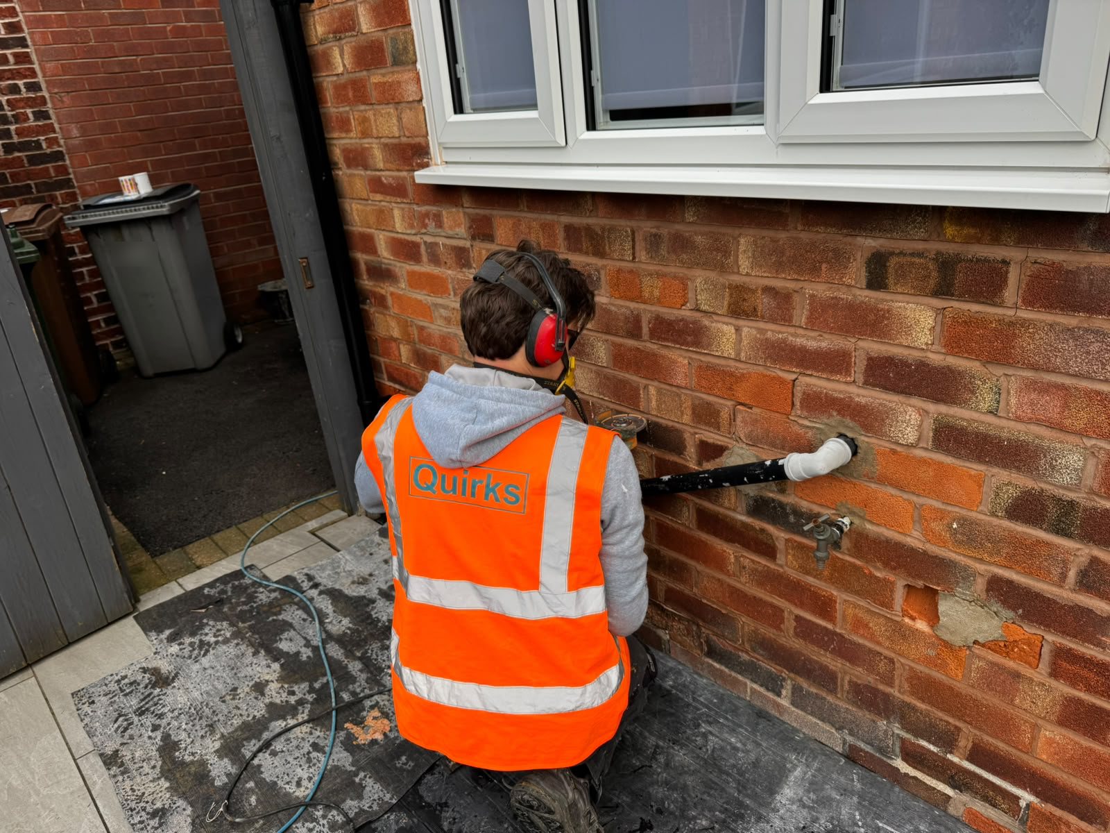 Operative in a Quirks hi-vis vest and ear defenders cutting out damaged bricks beside an outside tap and a black pipe
